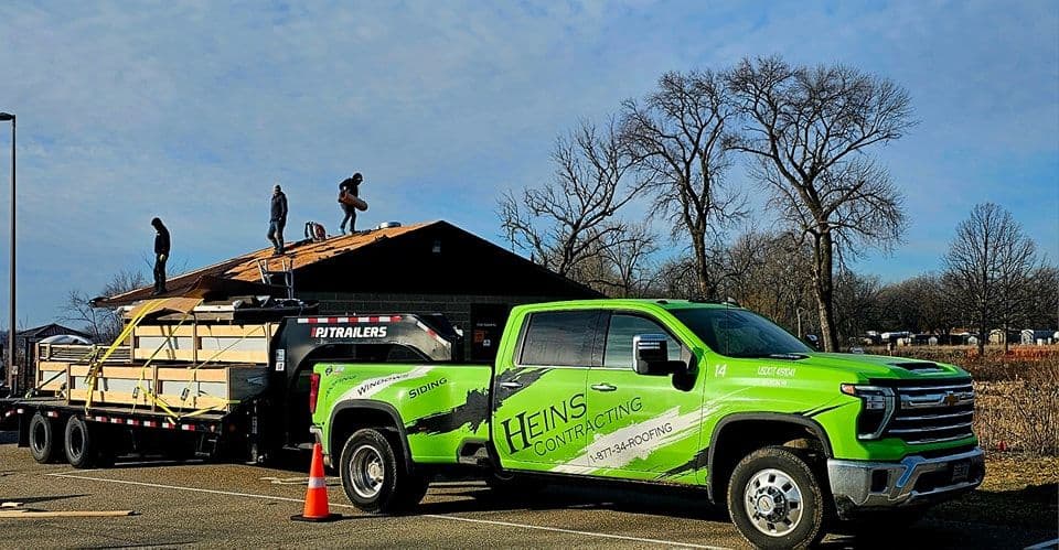 A Heins Contracting truck is parked in front of a house where roofers are working on installing a roof.